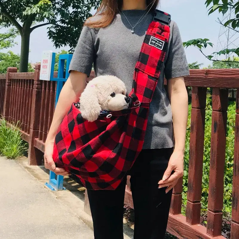 Woman carrying small white dog in red and black plaid pet carrier sling with adjustable shoulder strap, perfect for outdoor travel