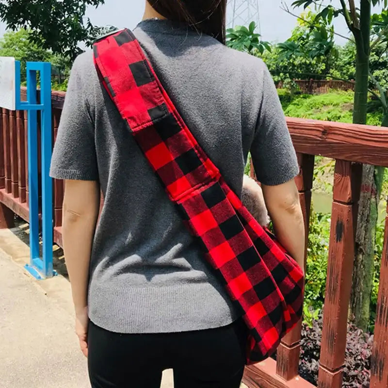 Person wearing a red and black checkered pet carrier sling outdoors on a wooden bridge.